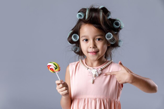 Funny little child girl in pink dress and hair curlers pointing lollipop on gray background.