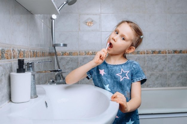 A funny little girl of 6 years old is standing in the bathroom near a white ceramic sink and carefully brushing her teeth