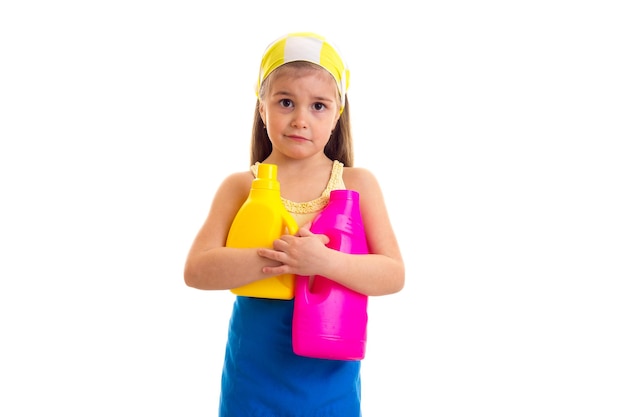 Funny little girl in blue apron with yellow kerchief holding yellow and pink bottles