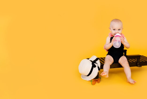 Смешная маленькая девочка, одетая в черно-розовый купальник, большая шляпа, сидит на деревянном шезлонге с бутылкой воды на желтом