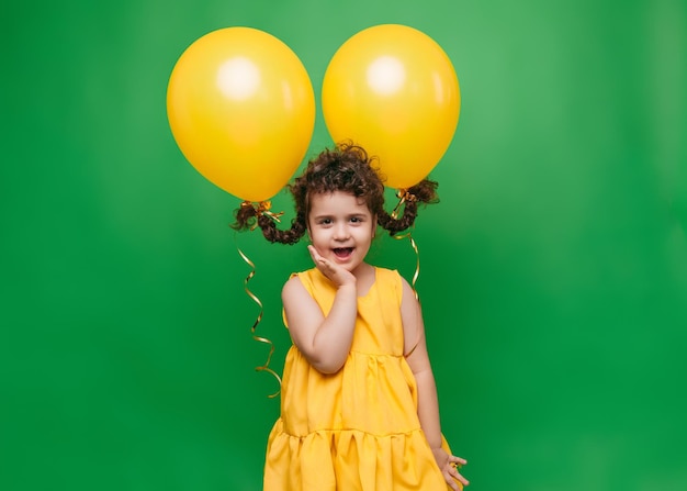 Funny little girl on a green background A girl is having fun with yellow balloons balloons lift up the pigtails of the child