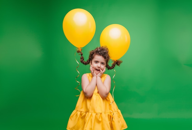 Funny little girl on a green background A girl is having fun with yellow balloons balloons lift up the pigtails of the child