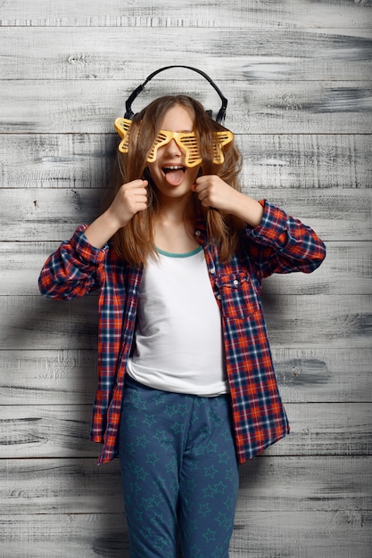 Photo funny little girl in headphones and star glasses in studio. children and gadget, kid isolated on wooden background, child emotion, schoolgirl photo session