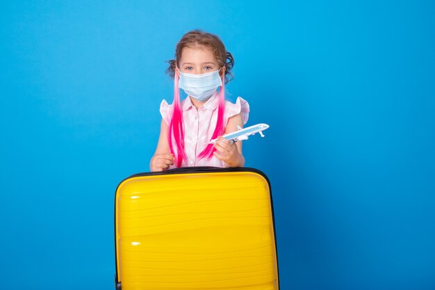 おもちゃの飛行機と青い表面に黄色のスーツケースと保護マスクの面白い女の子。