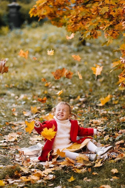 Funny little girl in a red coat catches fall leaves having fun and walking in the autumn forest