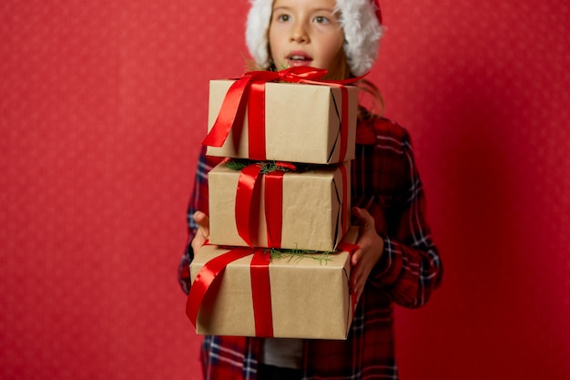 Bambina divertente in cappello della santa con molti contenitori di regalo di natale isolati su fondo rosso,