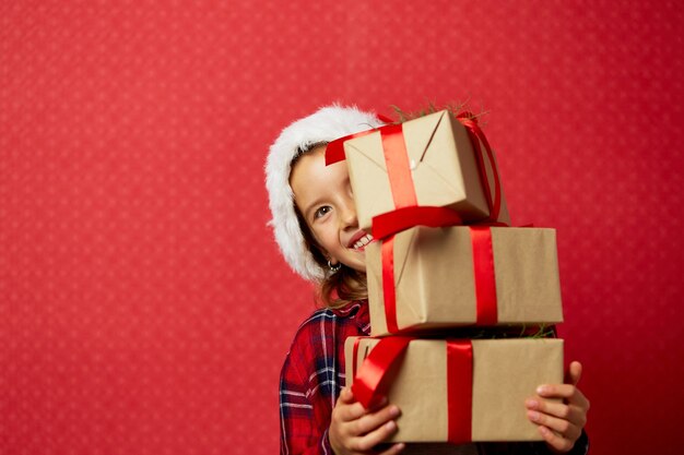 Funny little girl in santa hat with many Christmas gift boxes isolated on red background,