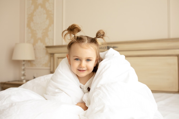 Photo a funny little girl sits on a big bed under a white fluffy blanket in the room