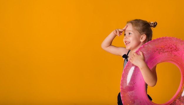 Una bambina divertente con un costume da bagno a righe tiene un cerchio gonfiabile rosa e guarda di lato su uno sfondo giallo con spazio per il testo