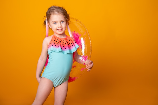 Funny little girl in swimming suit with swimming inflatable ring smiling having fun on yellow surface.