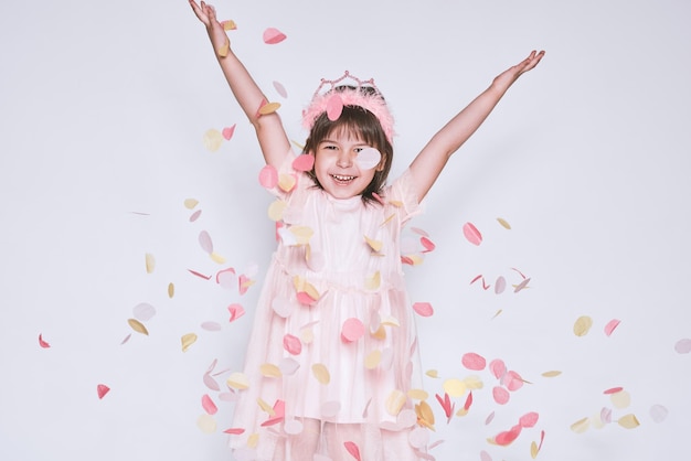 Funny little girl wearing pink dress in tulle with princess crown on head isolated on white background rise hands up enjoy confetti Pretty happy little girl celebrating her birthday party having fun