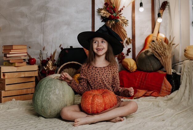 A funny little girl in a witch's hat is sitting on a knitted blanket with pumpkins