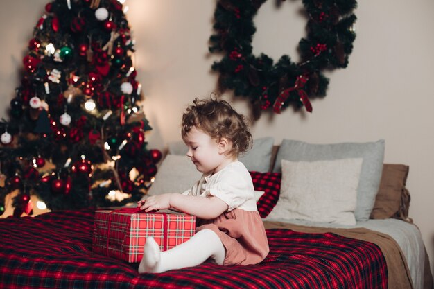 Funny little girl with curly hair sits on the bed near the Christmas tree with a present