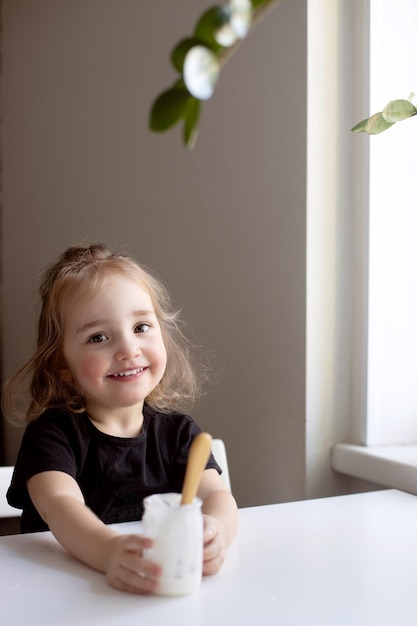 Foto bambina divertente con la faccia sporca che mangia yogurt fatto in casa da un barattolo di vetro usando un cucchiaio verticale