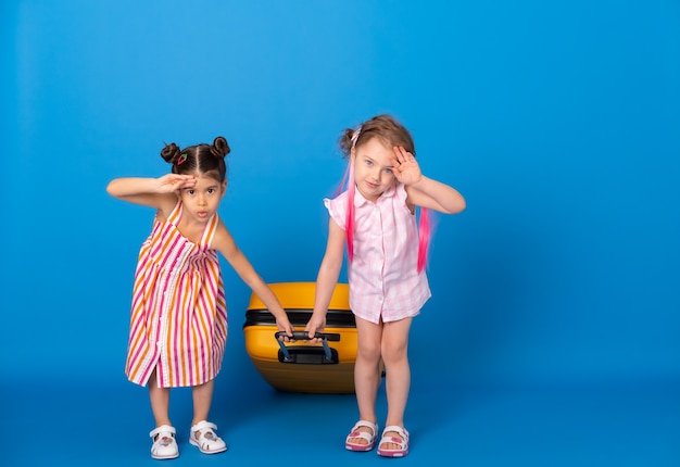 青い表面に黄色のスーツケースを運ぶカラフルな服を着た面白い小さな女の子の友達。