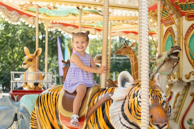 화려한 드레스를 입은 재미있는 어린 소녀는 여름날 놀이공원에서 회전목마를 탄다