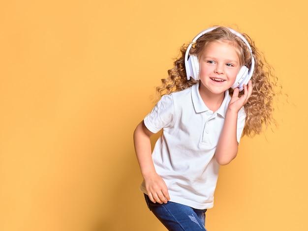 노란색 공간에 고립 된 흰색 티셔츠에 재미 있은 작은 꼬마 소녀. 어린 시절 라이프 스타일 개념. 복사 공간을 조롱하십시오. 펄럭이는 머리로 춤을 추며 헤드폰으로 음악 듣기