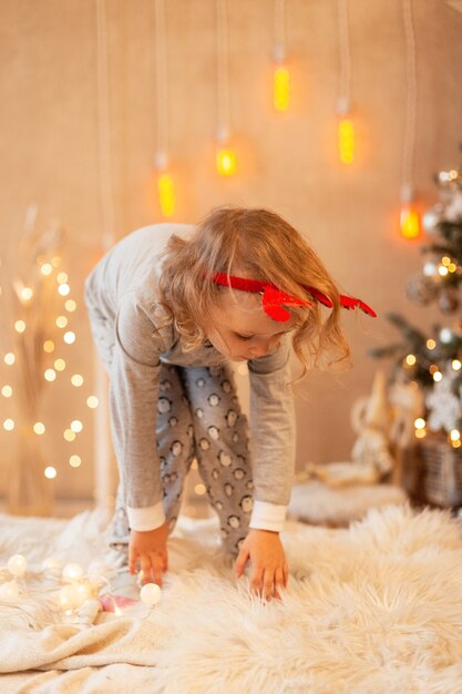 Funny little kid girl with horns jumping on the bed on a background of Christmas decorations and lights. Happy winter holidays