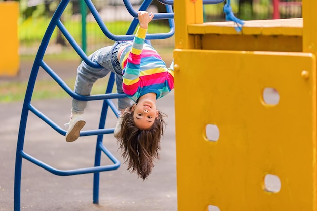 遊び場で子供の階段に逆さまにぶら下がっている面白い小さな笑顔の女の子