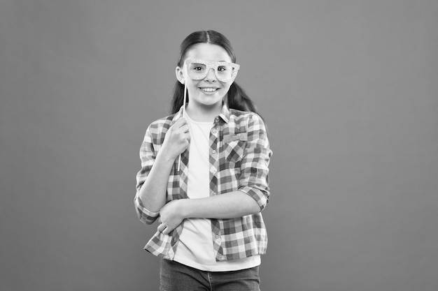 Funny look Adorable girl smiling with party look on orange background Cute little child having happy look through prop glasses Vogue look of small fashion model