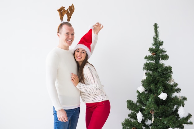 Funny loving couple near Christmas tree. Man wears deer horns and woman wears santa hat