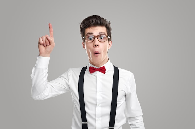 Funny male genius in glasses pointing up with forefinger while having brilliant idea and looking at camera on gray background