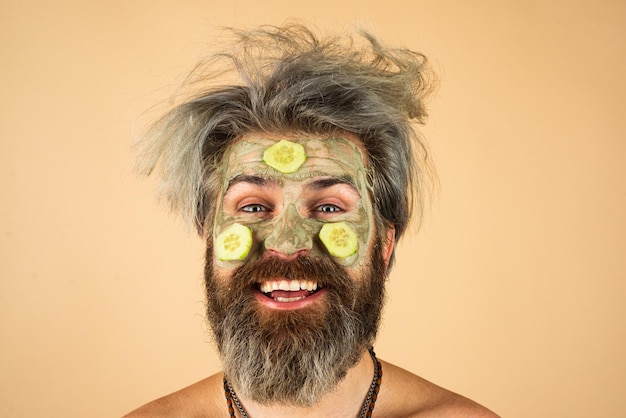 Funny man closeup with clay mask and cucumber slices on face
spa dermatology wellness and facial treatment concept man having
cosmetic moisturizing mask isolated studio background male face
care