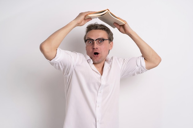 Funny man in glasses hiding under book holding it above head with amazement emotion on his face wear white shirt and grey jeans isolated white background learning concept