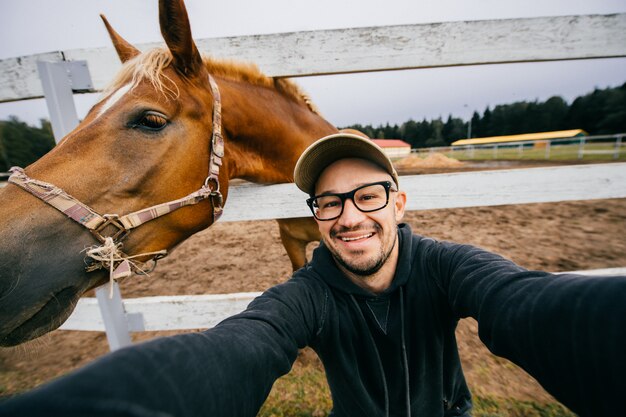 Забавный человек в очках принимая selfie с головой лошади за ним.