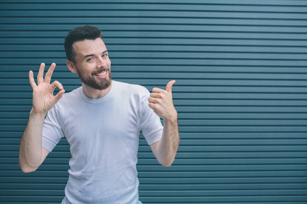 Funny man is showing ok and like symbols with his hands and fingers. Bearded man is looking on camera and smiling. Isolated on striped 