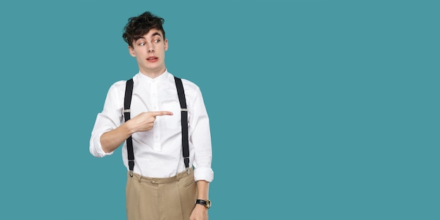 Funny man pointing and looking on empty copyspace. portrait of handsome hipster curly young businessman in classic casual white shirt, suspender standing. indoor studio shot isolated blue background.