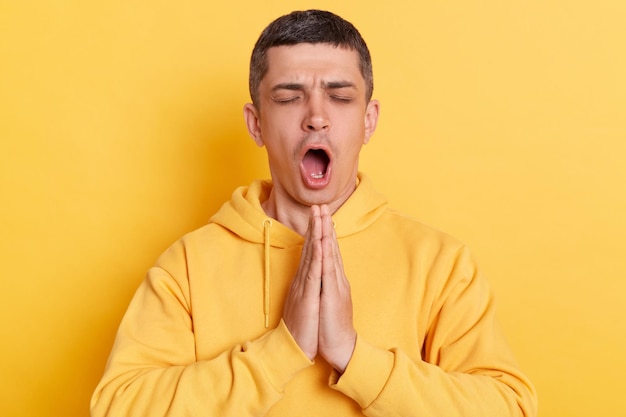 Funny man wearing casual hoodie meditating to calm down saying ohm standing with open mouth keeps palms together posing isolated over yellow background