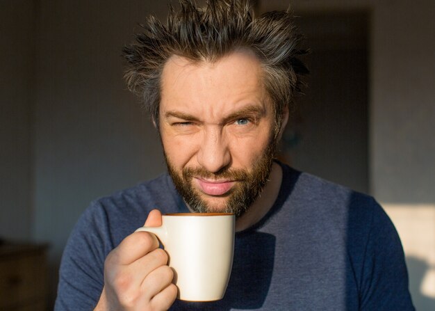 A funny man with a beard after waking up with a large cup of coffee or tea looks at the camera with ...