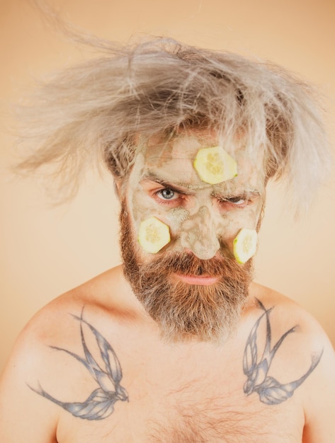 Funny man with clay mask and cucumber slices on face male spa dermatology wellness and facial treatm...