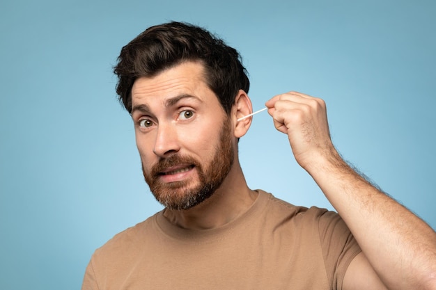 Funny middle aged bearded man cleaning ears with cotton swab doing daily hygiene routine blue studio background