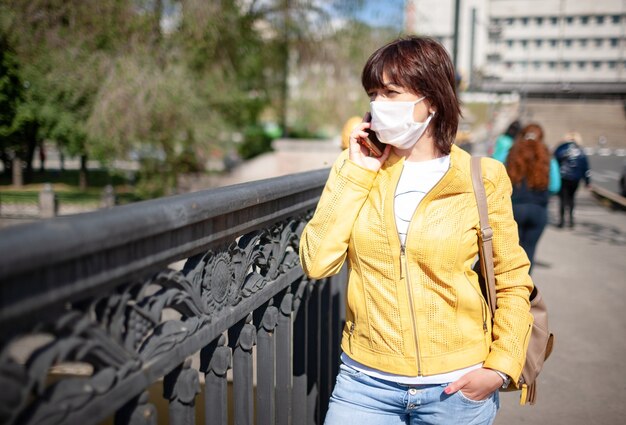 Funny middle-aged woman in white protective mask talking on smartphone while walking around city 