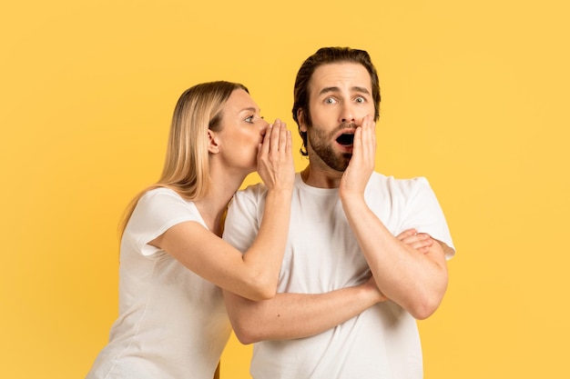 Funny millennial caucasian woman whispering in ear of surprised emotional man in white tshirt