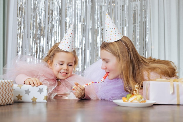 Funny moments of celebrating birthday of 5yearold girl Two sisters in party hats are happy together surrounded by gifts and cake with candle number 5 Happy childhood