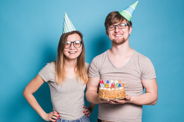 Funny nerd man and woman are wearing holidays caps and glasses holding birthday cake with candles over blue wall