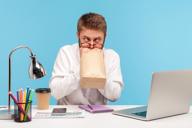 Funny nervous man office worker feeling sick rolling eyes\
breathing into paper bag to improve wellbeing overcoming stress at\
work indoor studio shot isolated on blue background