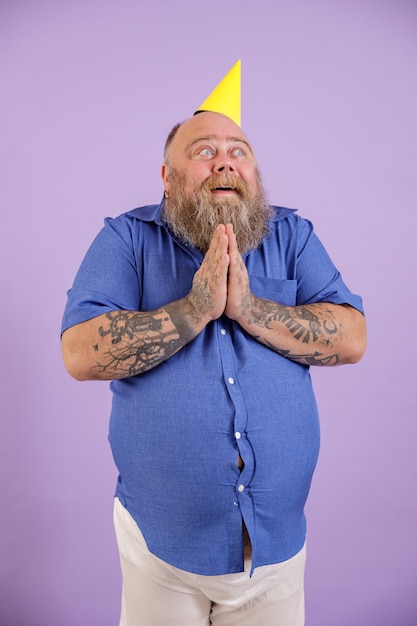 Funny obese male person holds hands in praying gesture on purple background