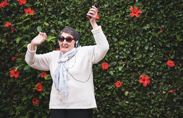 Photo funny old woman dancing listening to music on smartphone wearing earphones smiling enjoying fun celebrating retirement in garden with flower wall