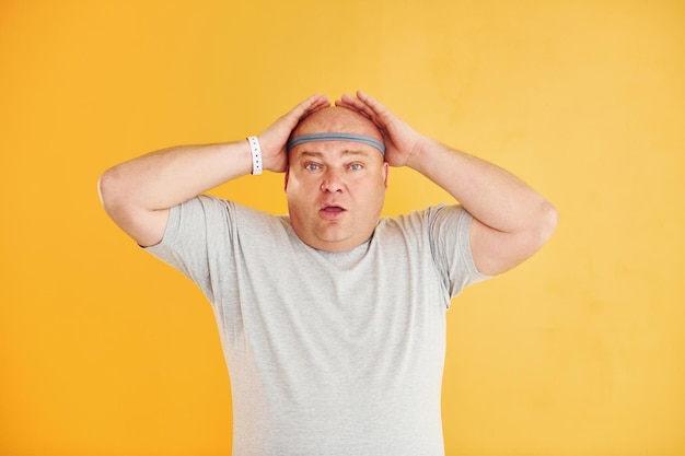 Funny overweight man in sportive head tie is against yellow background