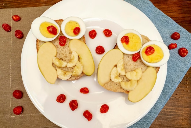 写真 面白いオオカミの顔のサンドイッチ トースト パン  ⁇ 卵 アップル バナナ 乾燥したベリー フルーツ 皿に 可愛い子供 子供の赤ちゃんの甘いデザート 朝食 ランチ 木製の背景の食品アートトップビュー