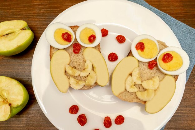 Funny owl face sandwich toast bread with chicken eggs apple banana dried berry fruits on plate Cute kids childrens baby's sweet dessert breakfastlunch food art on wooden backgroundtop view