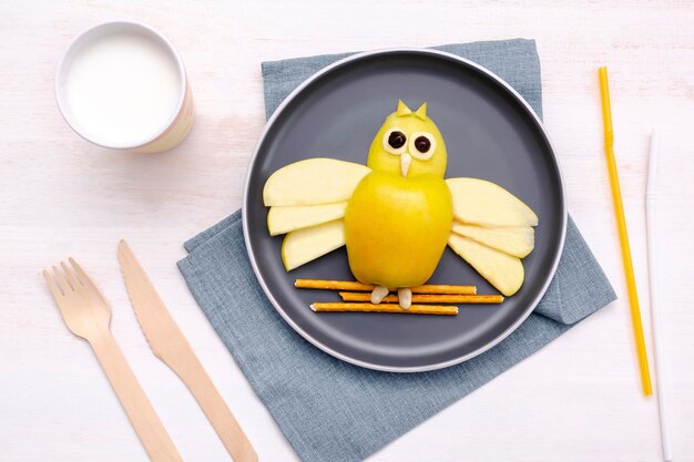 Funny owl face shape snack with apple wheat crispy bread straw on platemilk in glass Cute kids childrens baby's sweet dessert healthy breakfastlunch food art on wooden backgroundtop view