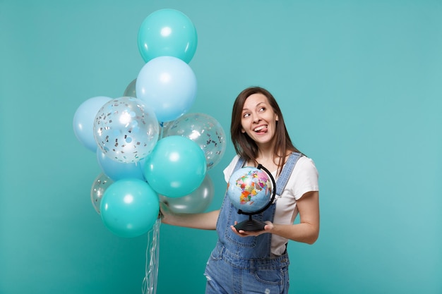 Funny pensive woman showing tongue, looking up holding Earth world globe, celebrating with colorful air balloons isolated on blue turquoise background. Birthday holiday party, people emotions concept.