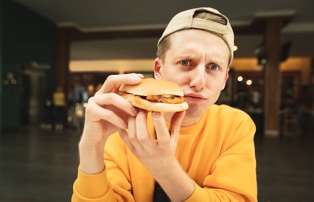 Фото Веселое фото веселого молодого человека в кепке и желтом свитере сидит в ресторане быстрого питания