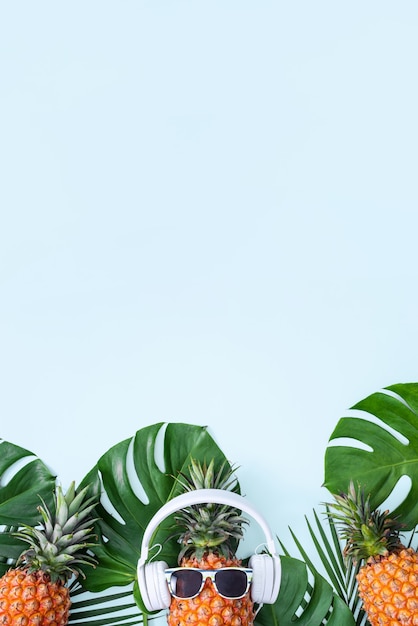 Ananas divertente che indossa cuffie bianche, concetto di musica d'ascolto, isolato su sfondo blu con foglie di palma tropicale, vista dall'alto, design piatto.