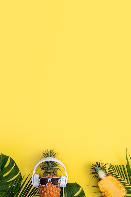 Funny pineapple wearing white headphone listen music isolated on yellow background with tropical palm leaves top view flat lay design concept